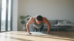 Mann macht Liegestütze im Wohnzimmer auf einer Fitnessmatte, Sonnenlicht fällt herein.